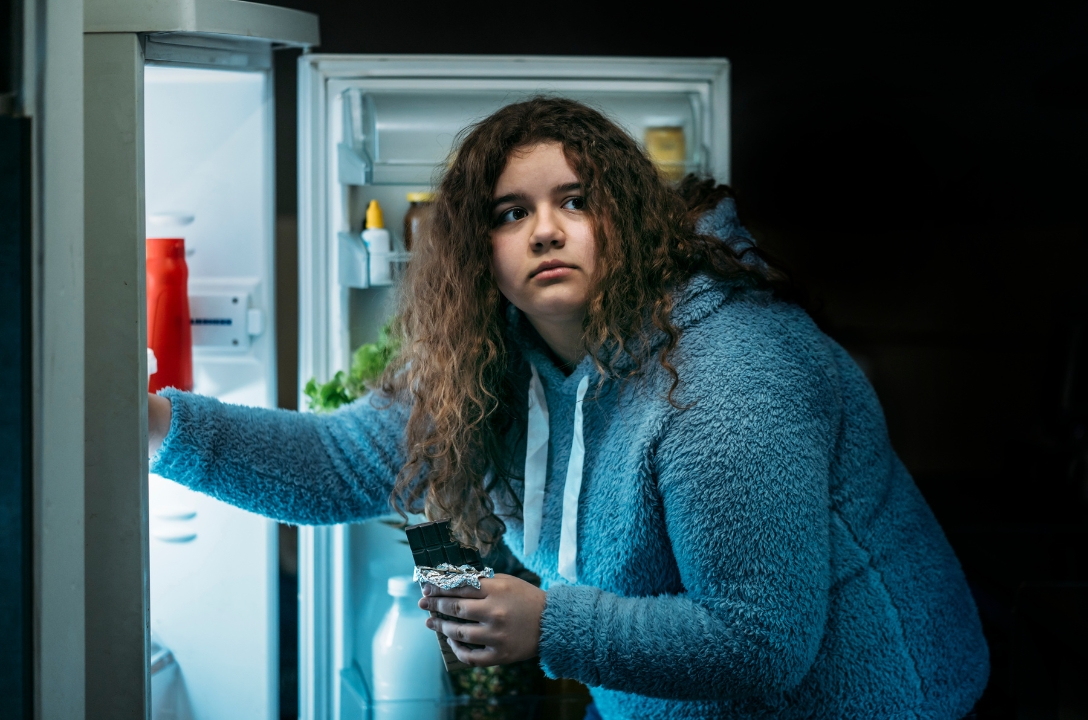 Girl in a grey jumper goes to the fridge to get some chocolate and eat it in secret