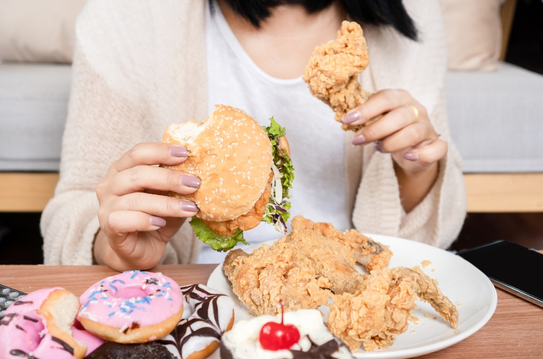 woman holding a burger in one hand and a chicken drumstick in the other hand. Pink doughnut and cupcake are on the table
