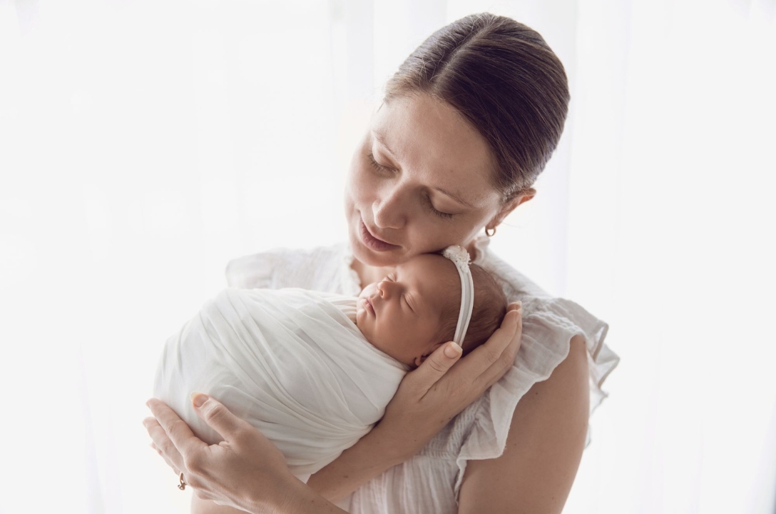 Woman in a white dress lovingly holding a new born baby with a white headband against a white background